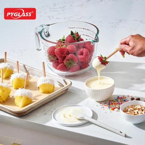 Hand dipping a strawberry into white chocolate, surrounded by a measuring cup of raspberries, pineapples, sprinkles, and almonds.