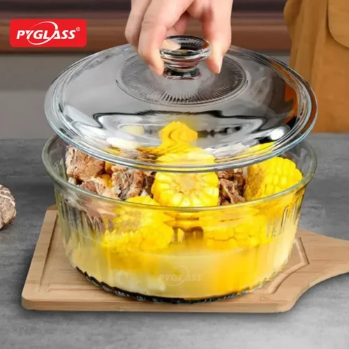 A clear glass casserole dish with a lid partially lifted, containing yellow corn and meat on a wooden board.