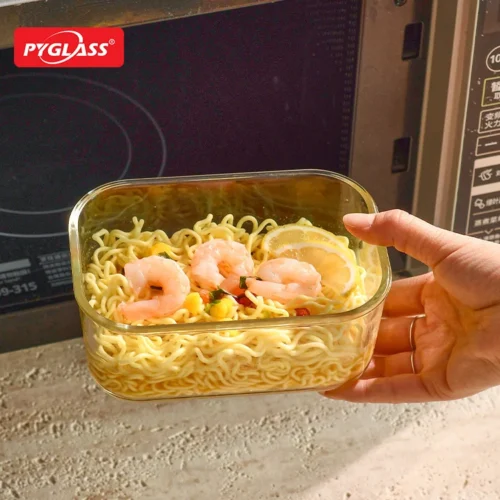 A hand holding a glass container with shrimp noodles, garnished with lemon slices, near a microwave on a kitchen counter.