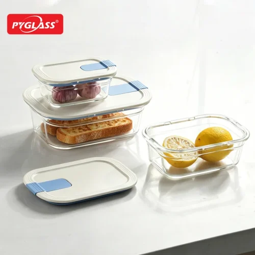 Set of glass food storage containers with blue-tinted lids, containing bread, lemons, and garlic on a white surface.