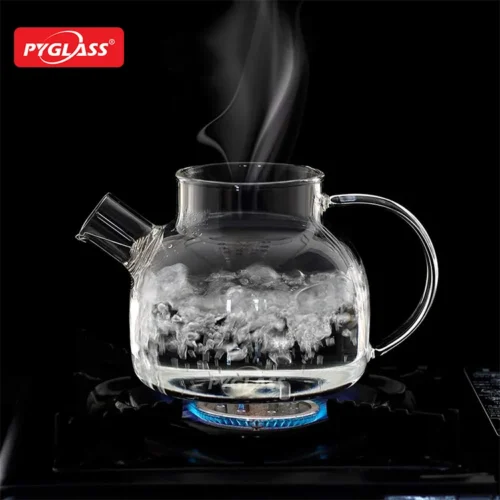 Clear glass teapot with boiling water, releasing steam from spout on a gas stove with blue flame.