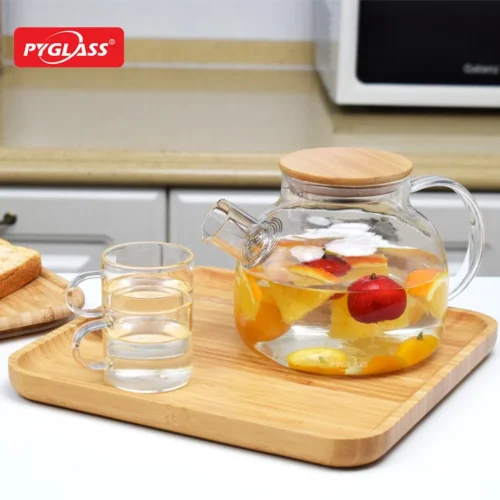 Glass teapot with a wooden lid, filled with fruit-infused water, placed on a wooden tray with two glass cups and toast in a kitchen setting.