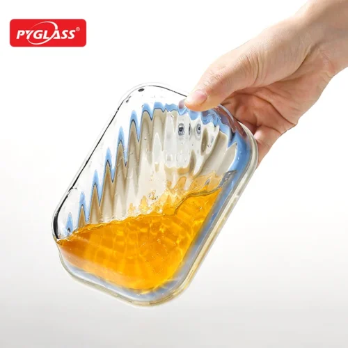 A hand holding a clear glass dish partially filled with orange liquid, shown against a white background.