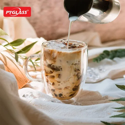 Glass of iced coffee with milk being poured from a jug, placed on a fabric surface surrounded by leaves and natural decor.