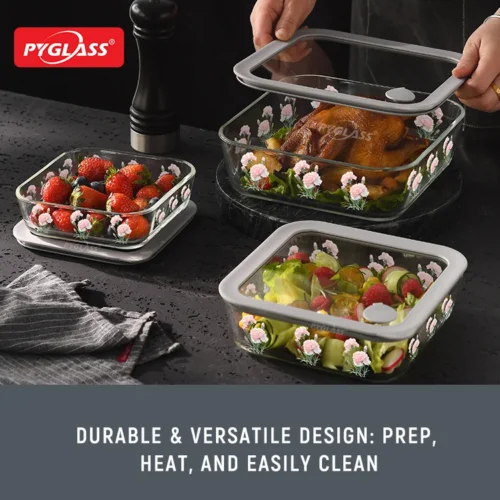 Three Pyglass glass containers with floral designs holding strawberries, a salad, and a roasted dish. One container is being uncovered by hand.