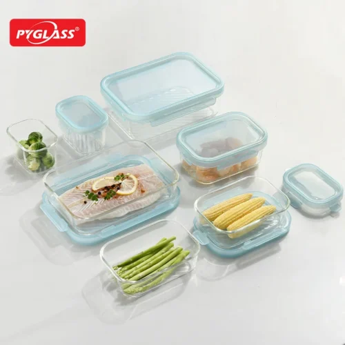 A variety of Pyglass food storage containers holding fresh foods like asparagus, fish, brussels sprouts, and corn on a white surface.