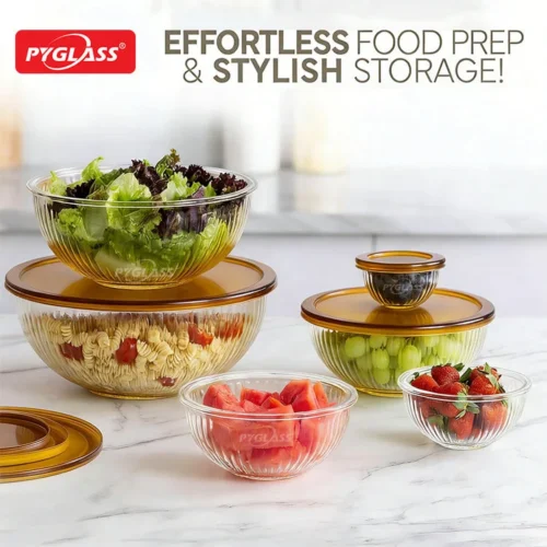 Assorted Pyglass glass bowls containing fresh vegetables and fruit with amber lids on a marble countertop.