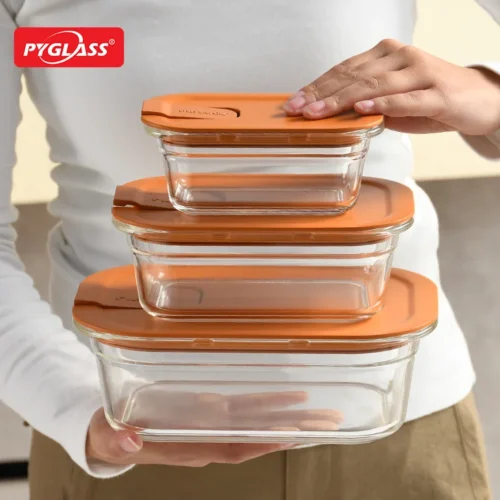 A person holding a stack of three glass food storage containers with orange lids, showcasing a practical kitchen organization solution.
