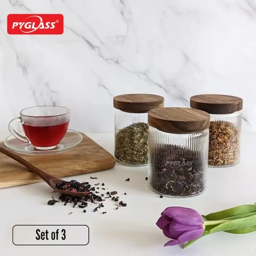 Three glass jars with wooden lids filled with various teas. A glass cup of red tea sits beside them on a wooden board. A purple tulip in the foreground.